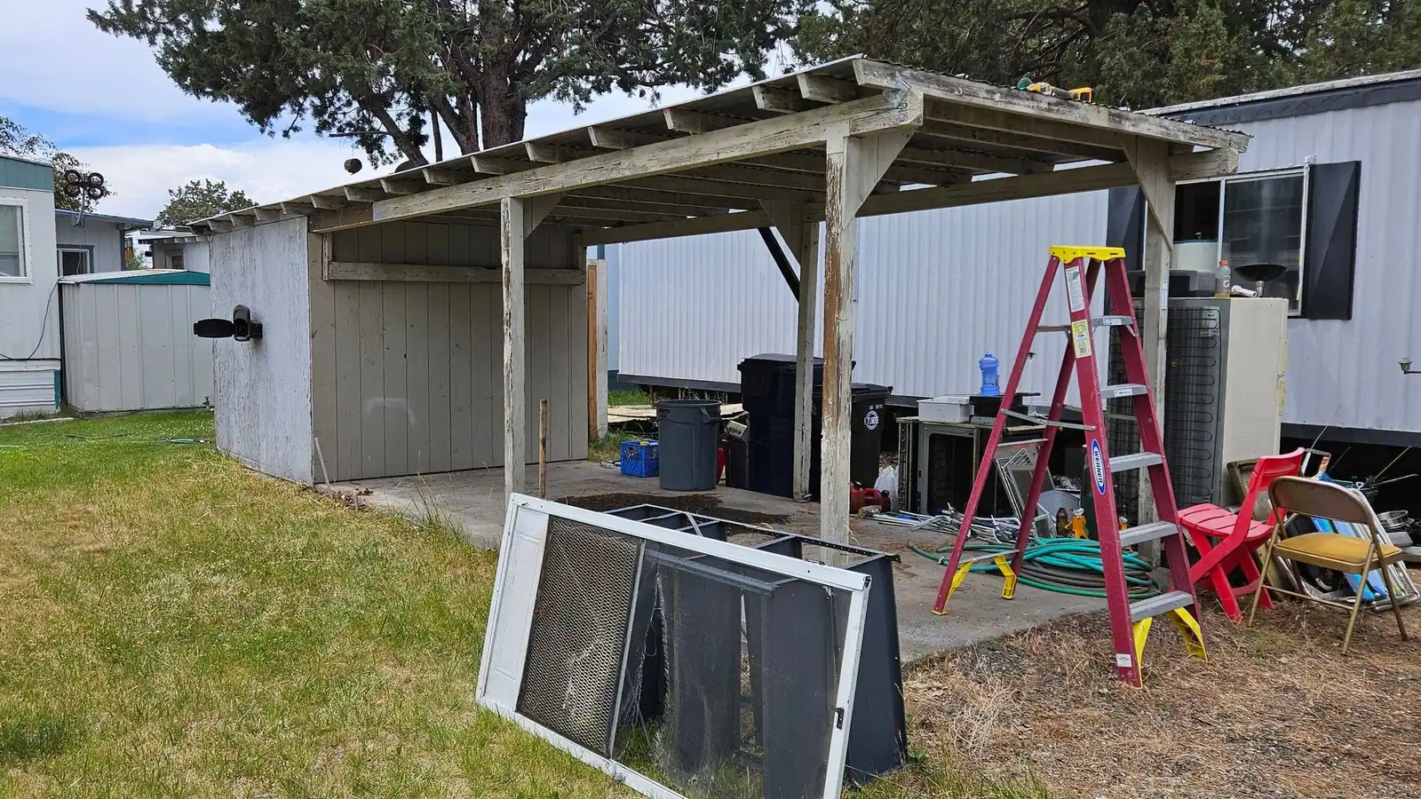 A carport with an old screen door, a ladder, hoses and bins waiting to be cleared