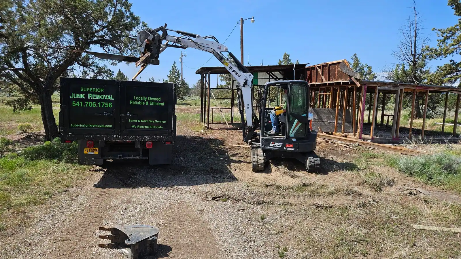 A mini excavator tearing down an old wooden structure with the Superior Junk Removal truck parked beside it