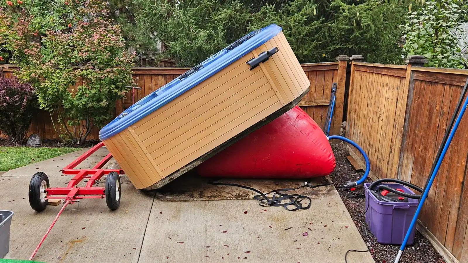 An old hot tub tipped up on a red inflatable lift beside a trailer on a deck
