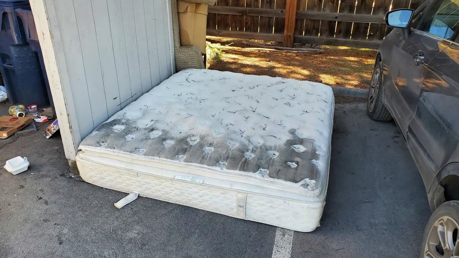 An old stained mattress against a fence in a parking spot