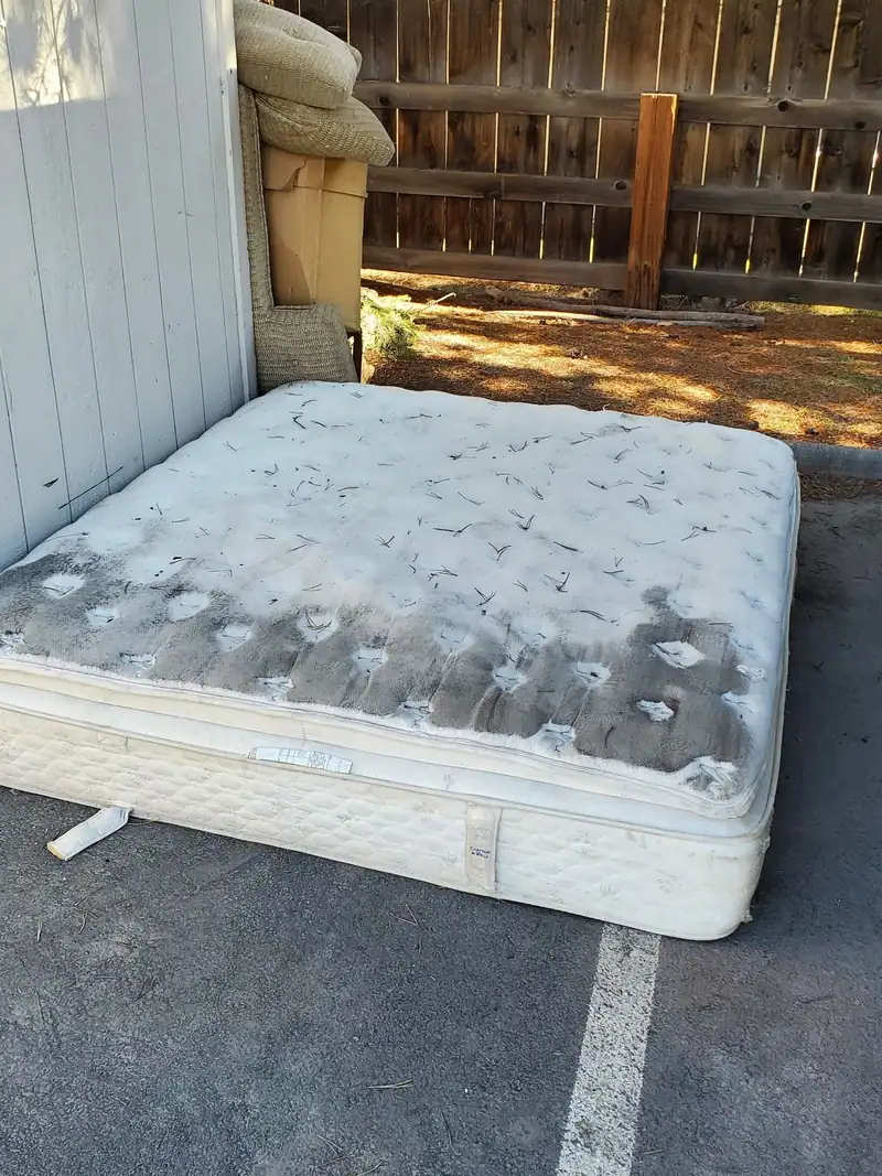 An old stained mattress against a fence in a parking spot