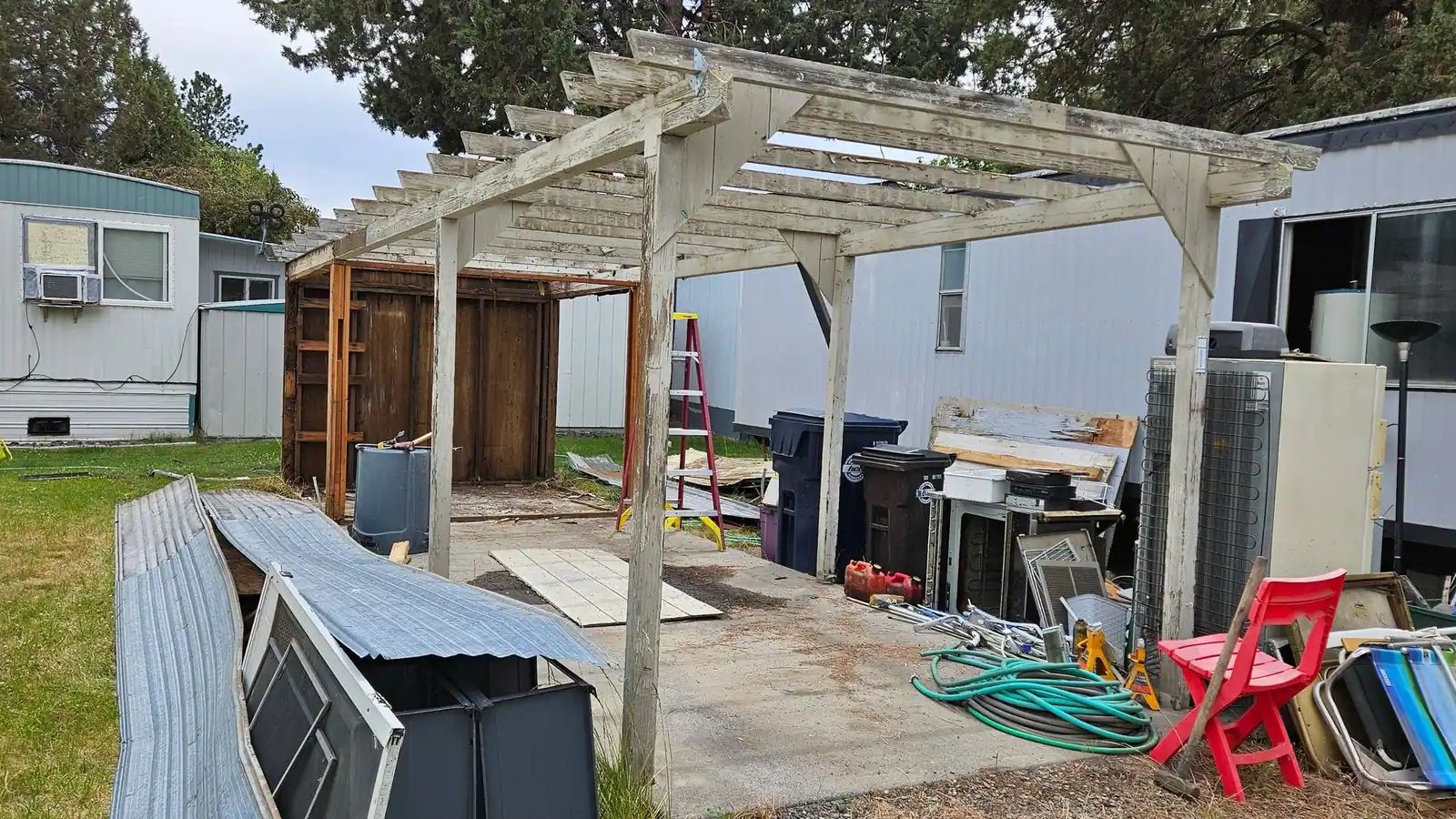 A backyard under a pergola with an old grill, hoses, bins and scrap waiting to go