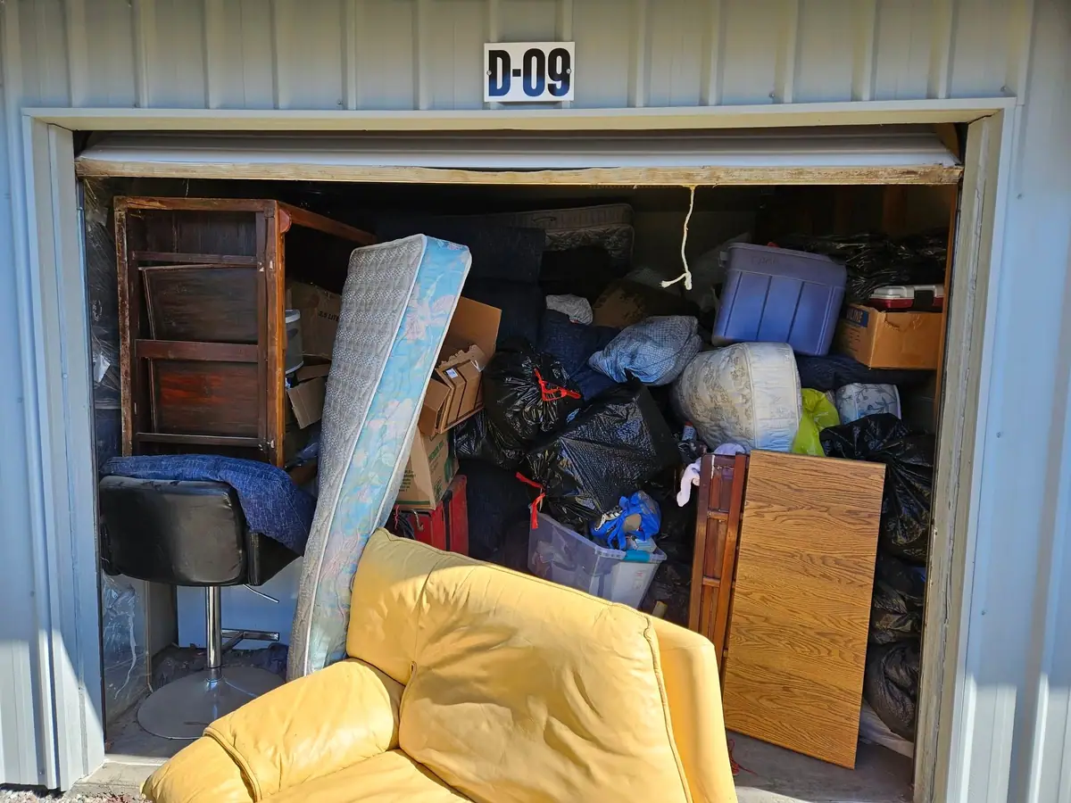 A storage unit packed to the door with furniture, a mattress, bags and a yellow armchair
